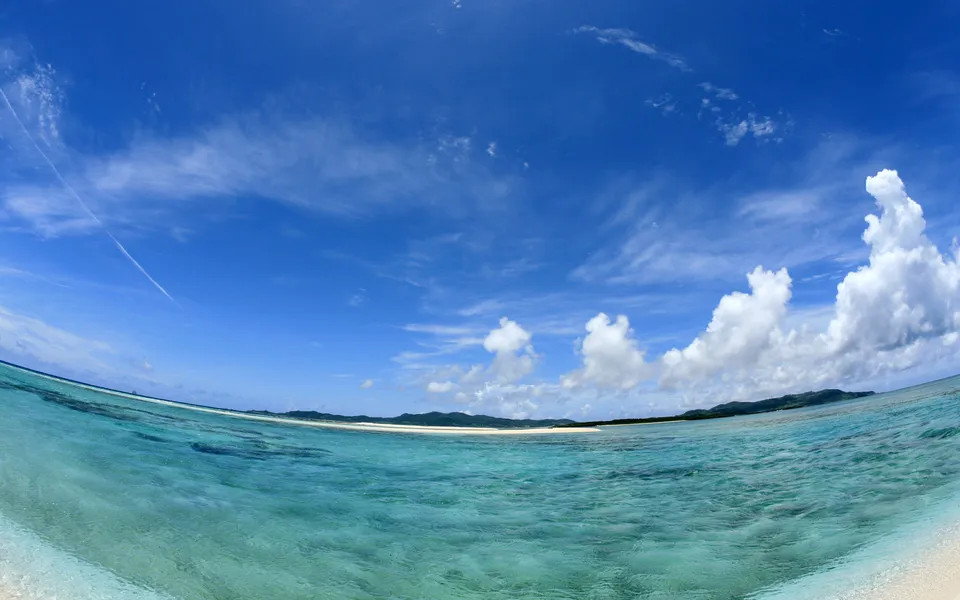 A wide-angle view of a tropical beach with clear turquoise water and a blue sky with scattered clouds.