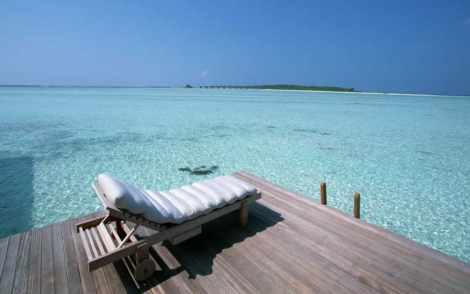 A lounge chair sits on a wooden deck overlooking crystal clear turquoise water and a distant island.