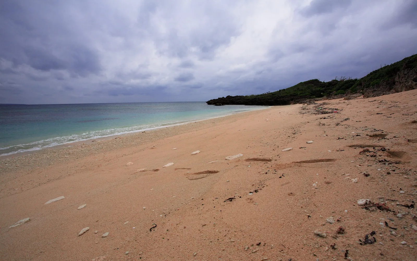 Footprints mark a sandy beach leading to the turquoise ocean under a dramatic cloudy sky.