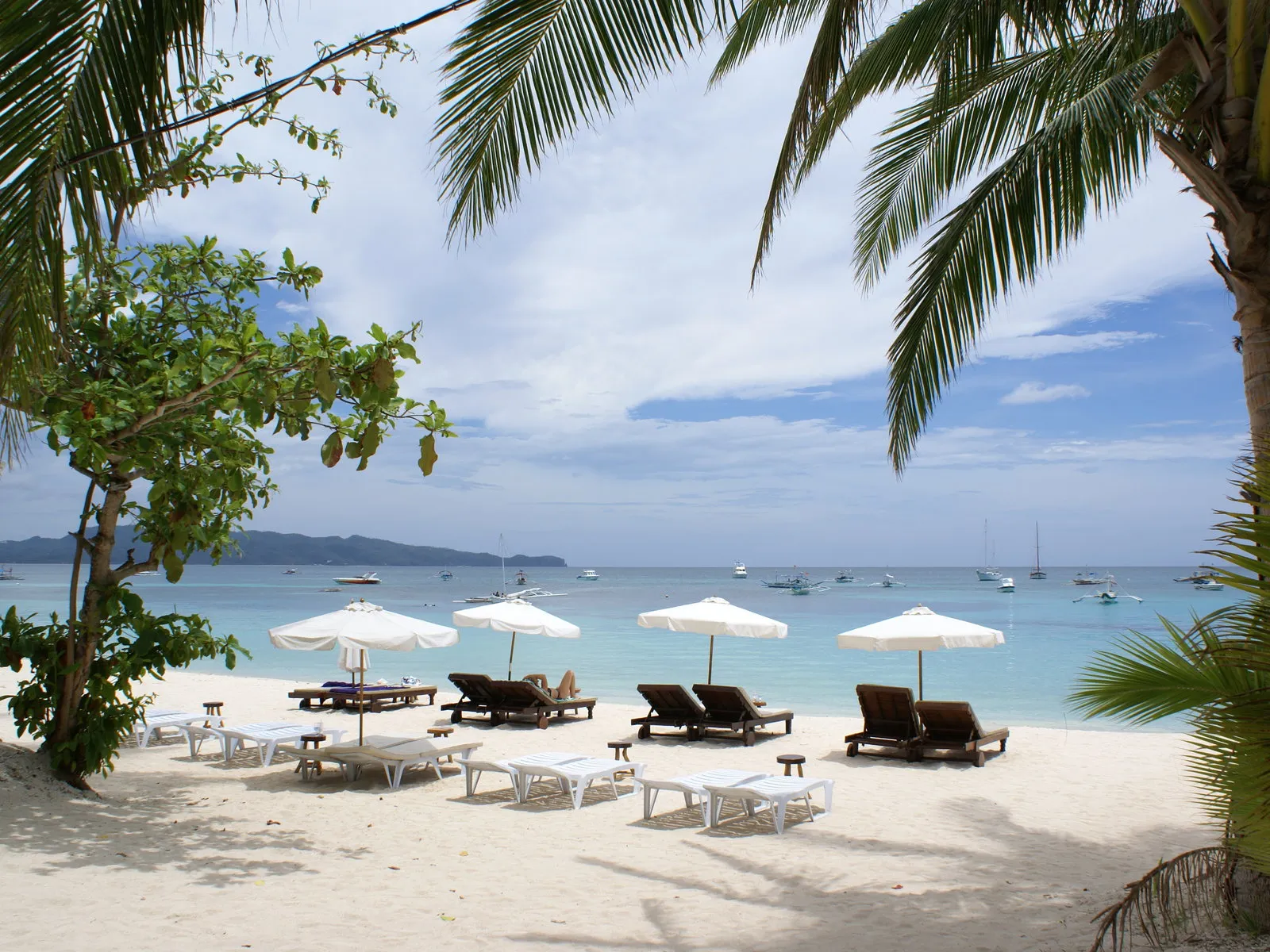 A tropical beach with numerous lounge chairs and umbrellas, with many boats on the horizon.