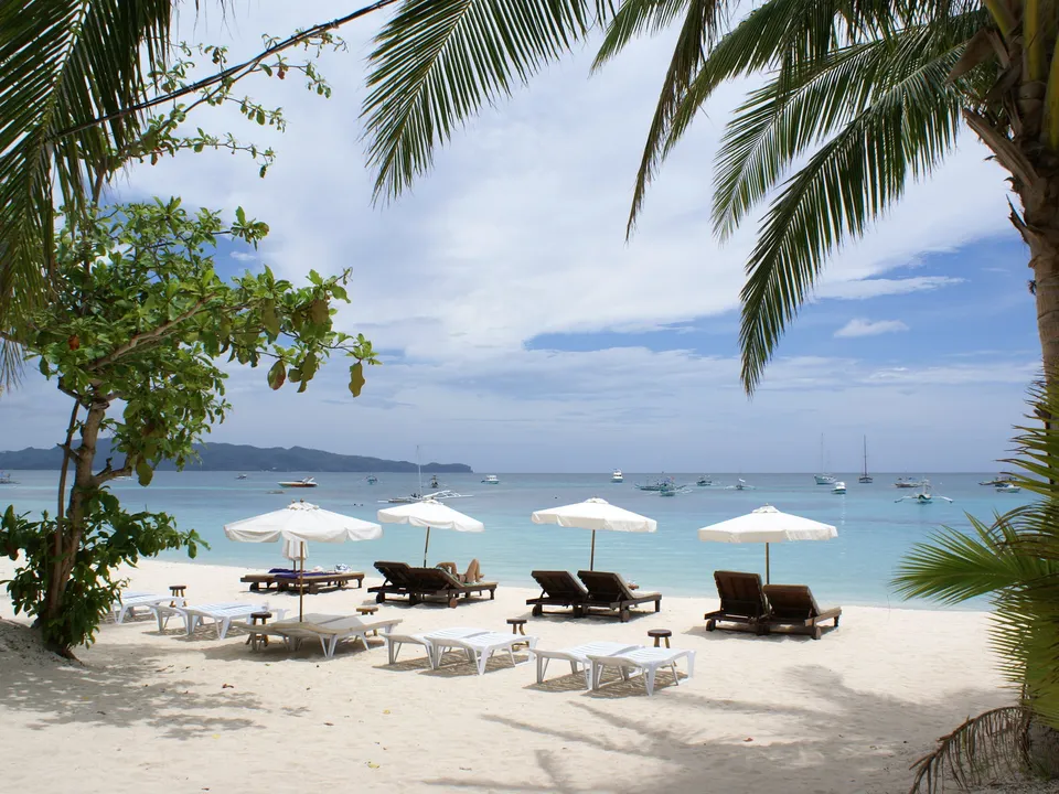 A tropical beach with numerous lounge chairs and umbrellas, with many boats on the horizon.