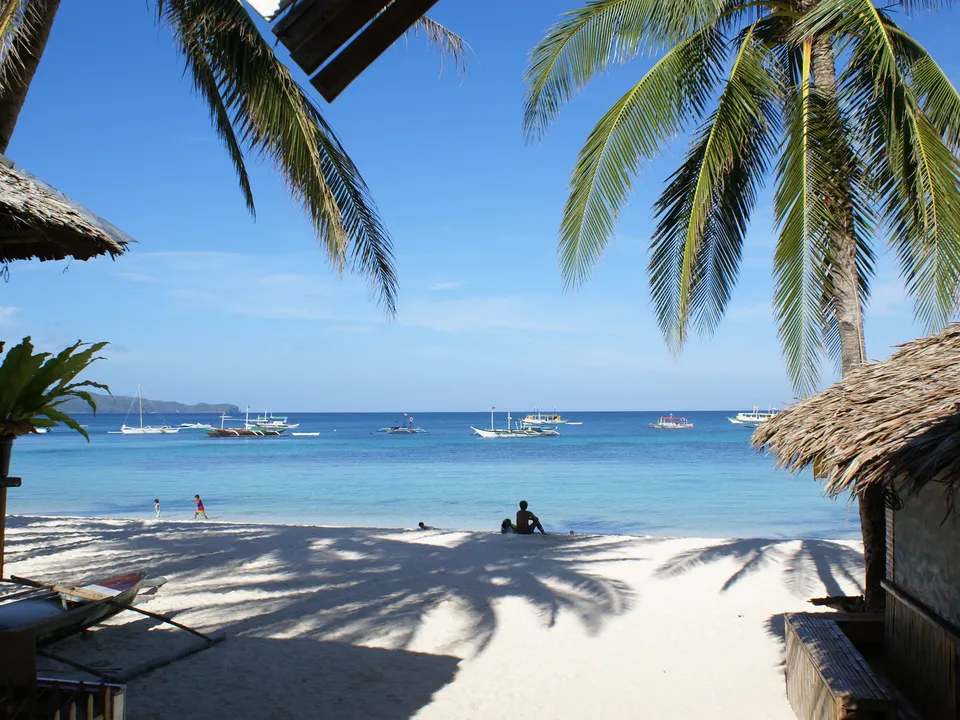 View of a tropical beach with palm trees, white sand, and boats in the turquoise ocean.