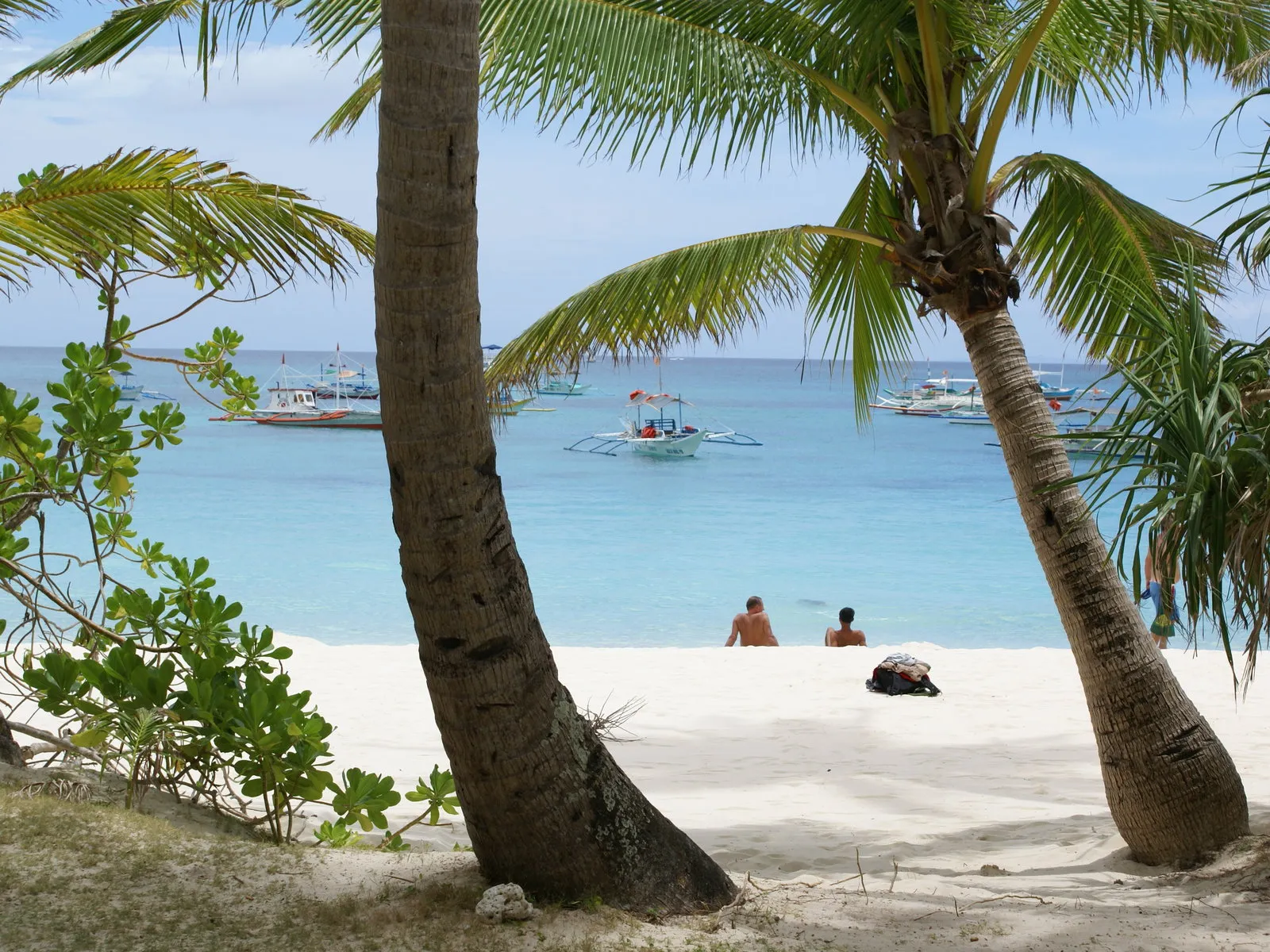Two people sit on a white sand beach with palm trees in the foreground and boats in the turquoise ocean.