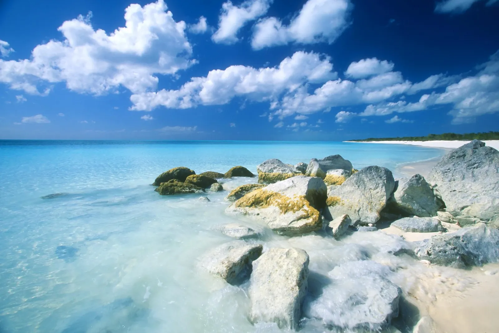 A view of a tropical beach with clear turquoise water, white sand, and large rocks in the foreground.
