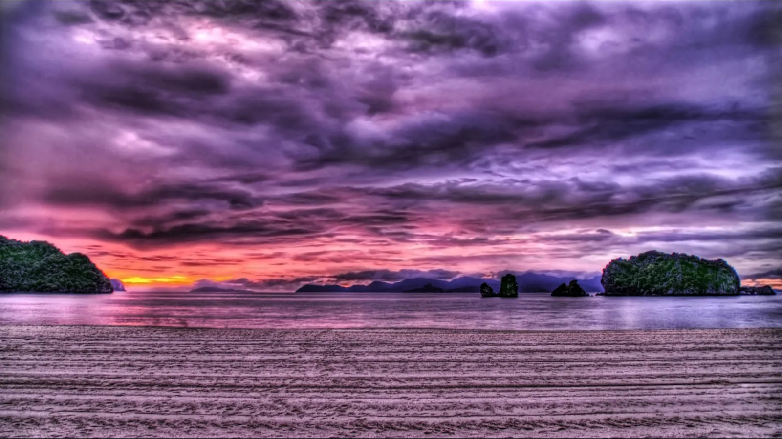 A dramatic sunset with purple and pink clouds over a tropical ocean with islands and a sandy beach in the foreground.