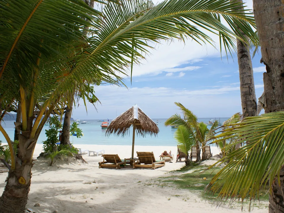 A tropical beach with palm trees, a thatched umbrella, and lounge chairs facing the ocean.