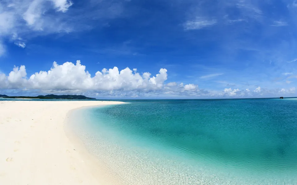 A panoramic view of a white sand beach meeting clear turquoise ocean water under a blue sky with white clouds.