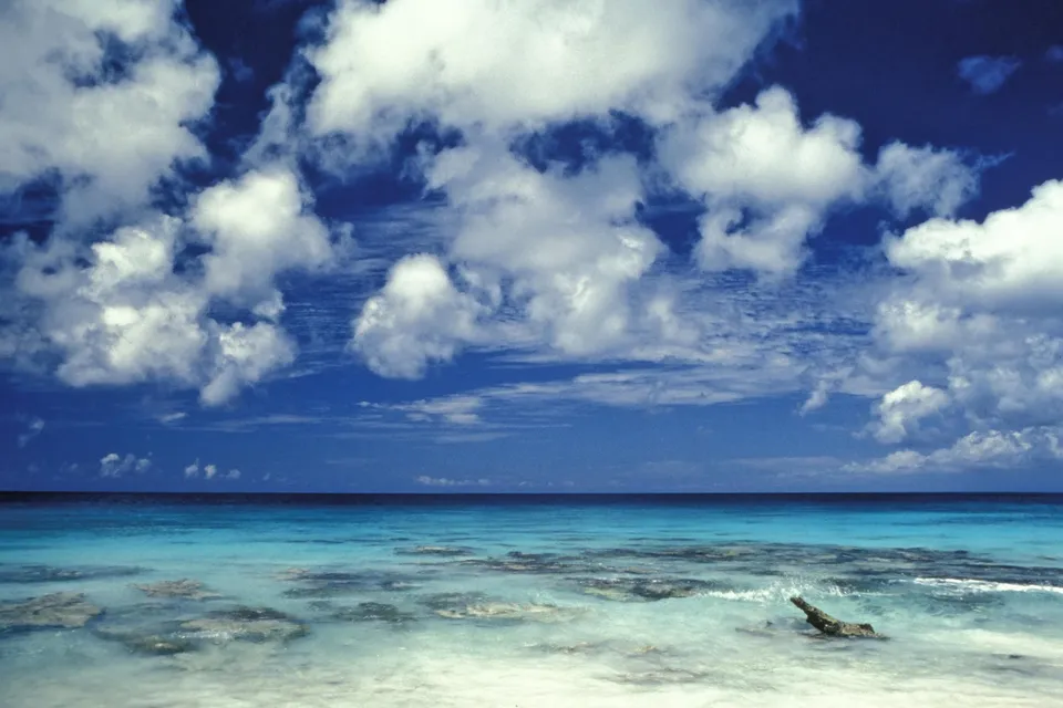 A view of a tropical beach with clear turquoise water, shallow coral formations, and a bright blue sky with fluffy white clouds.