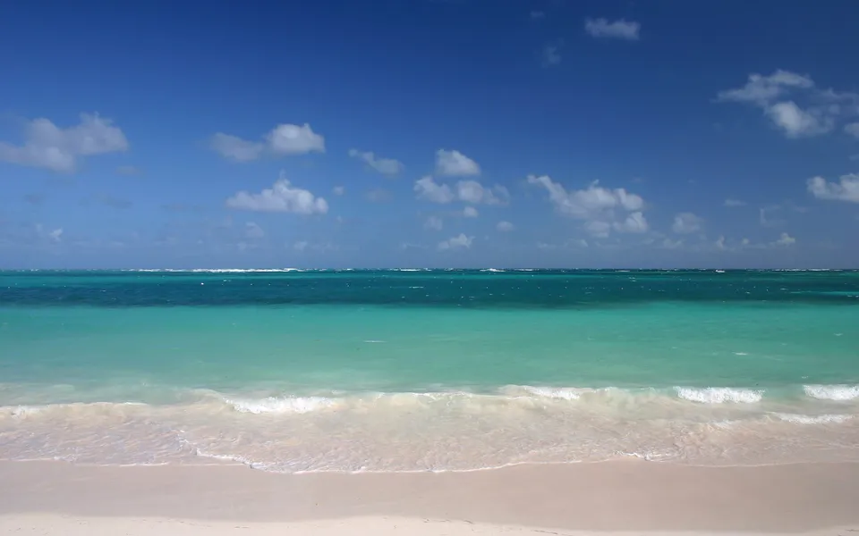 A serene tropical beach with white sand, gentle waves, and clear turquoise ocean water under a bright blue sky with scattered clouds.