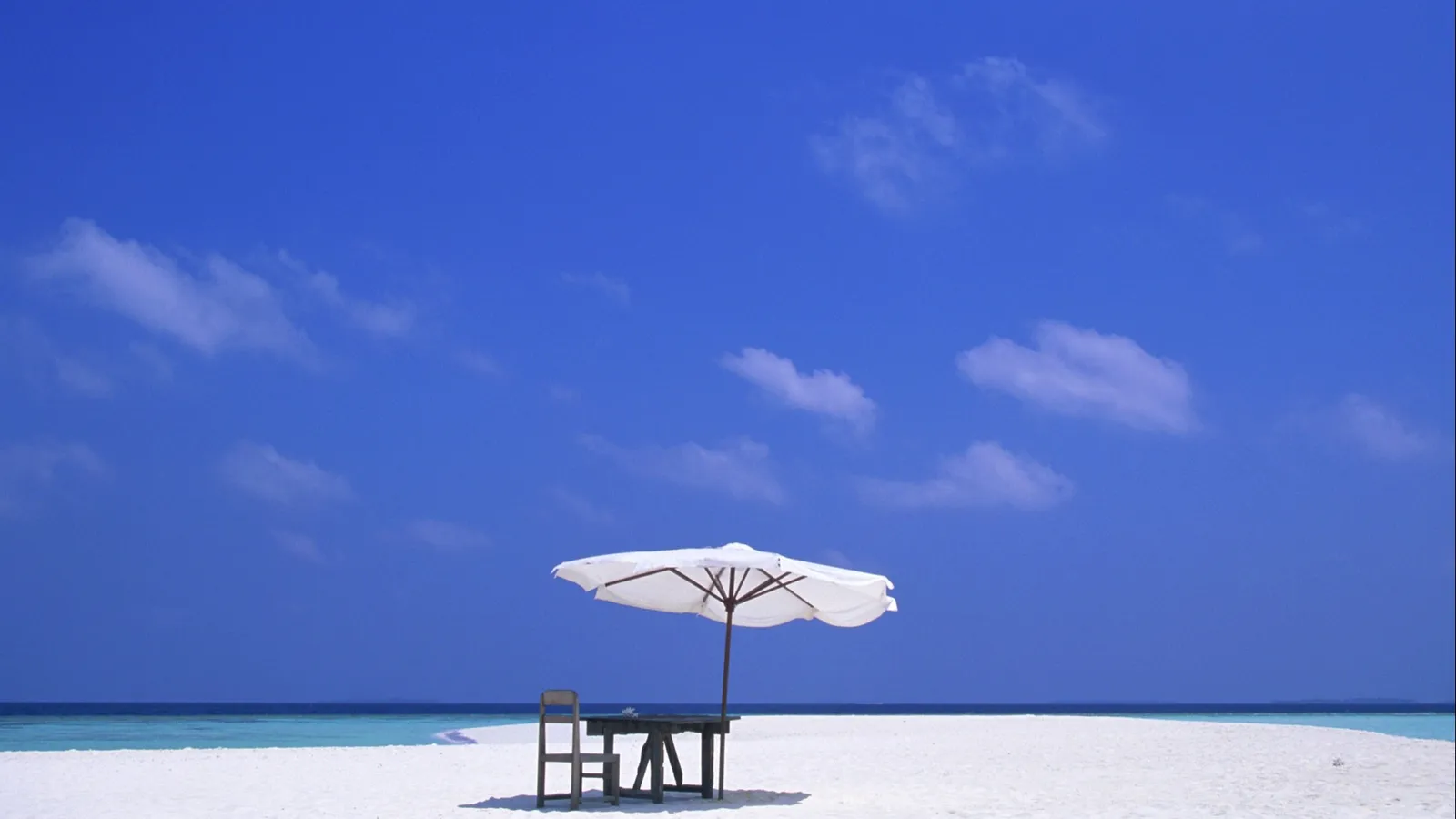 A white umbrella and a chair sit on a white sand beach under a bright blue sky with a few clouds.