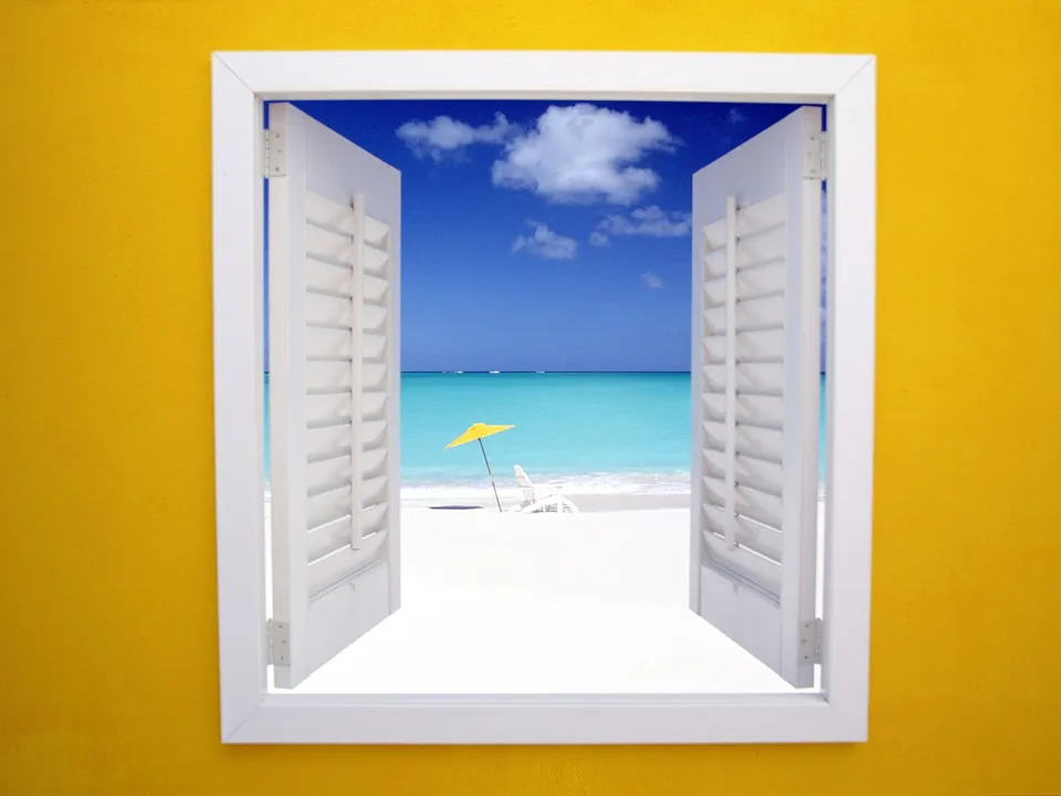 View of a bright white sand beach with turquoise water and a yellow umbrella through an open window with white shutters.