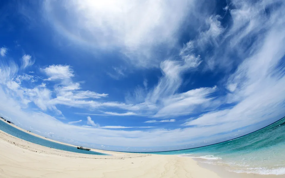 A wide sandy beach meets turquoise water under a bright blue sky with scattered wispy white clouds.