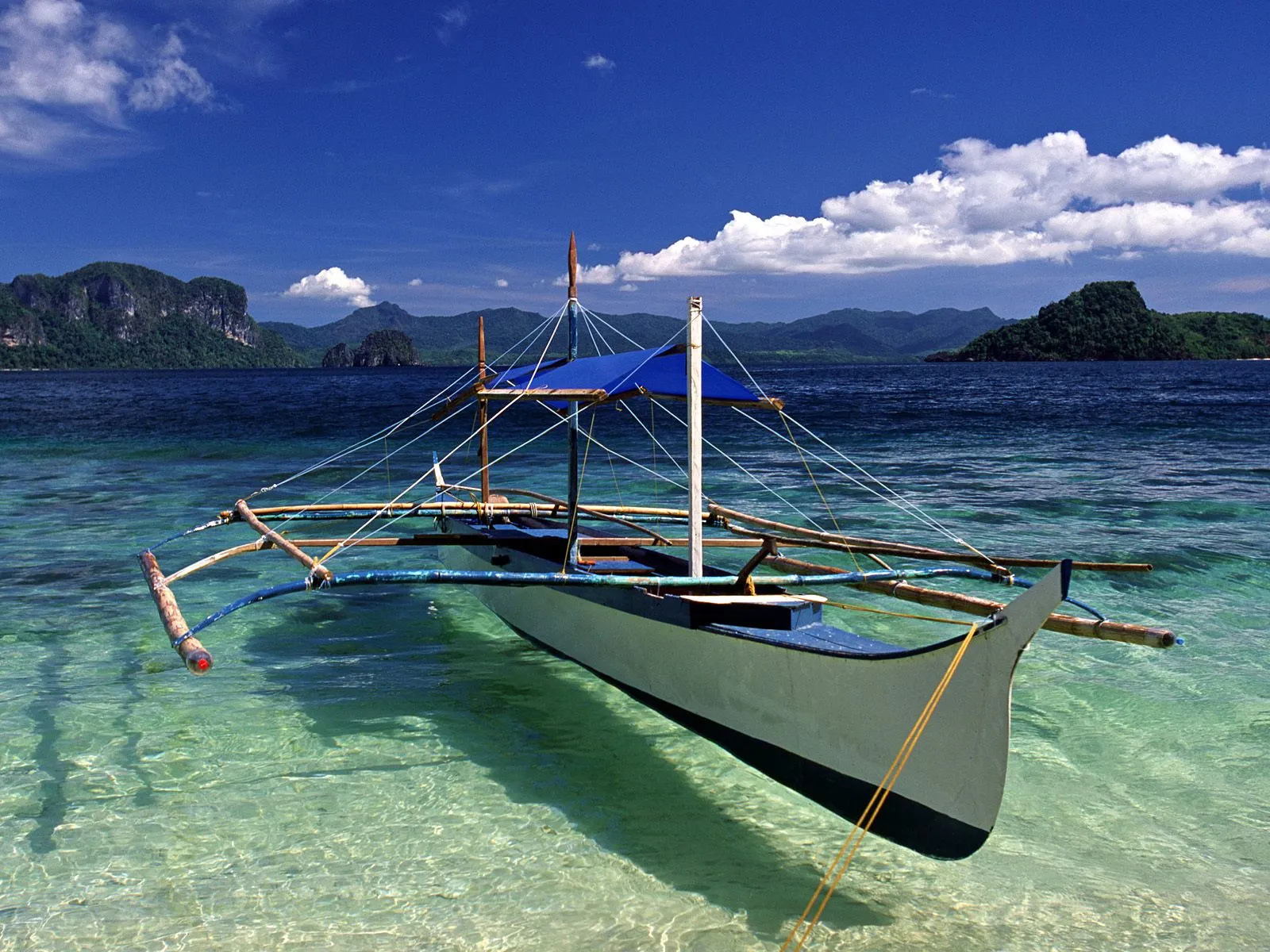 A traditional Filipino banca boat floats in shallow, clear turquoise water near a tropical island with lush green hills.