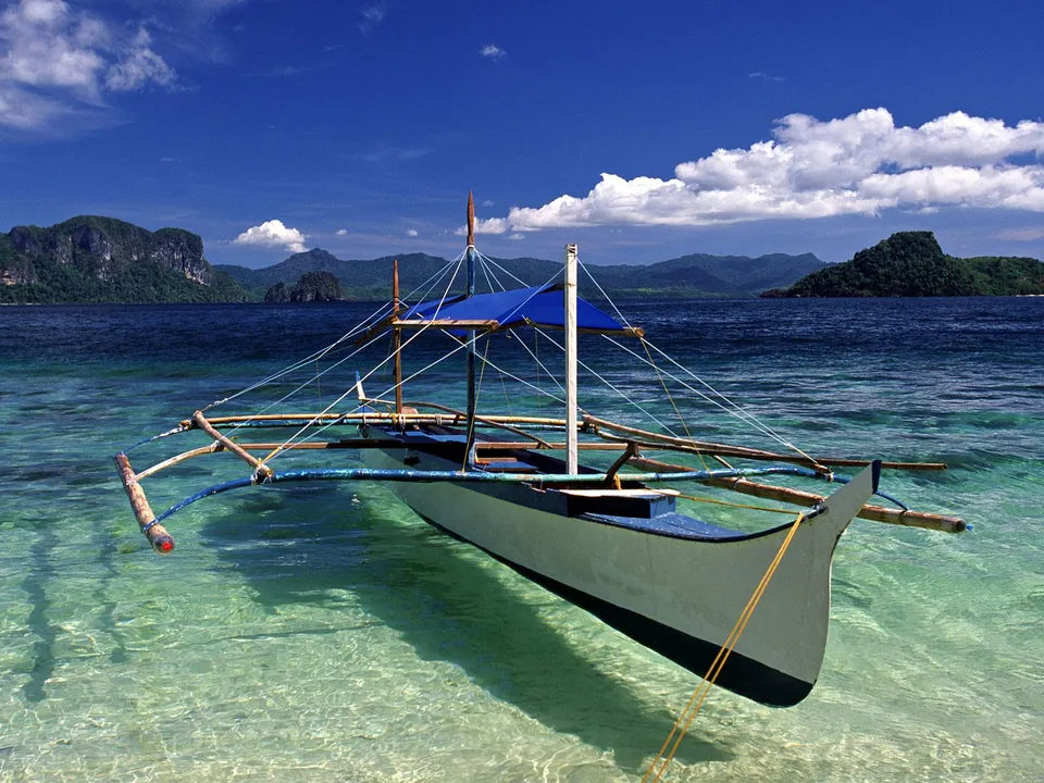A traditional Filipino banca boat floats in shallow, clear turquoise water near a tropical island with lush green hills.