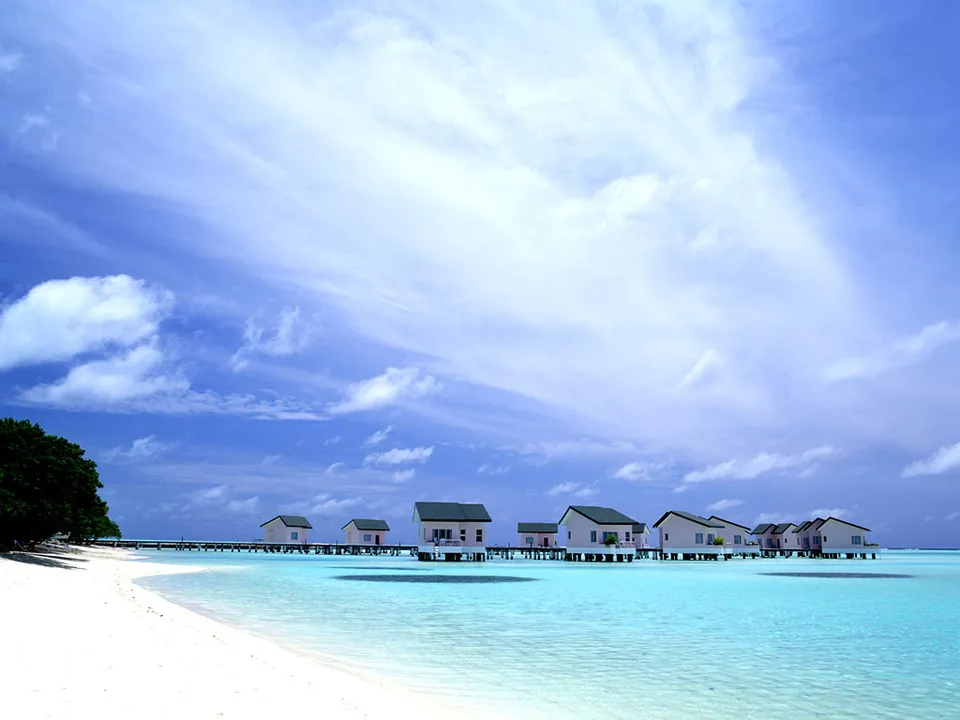 Overwater bungalows line a pier extending into turquoise water, with a white sand beach and blue sky.
