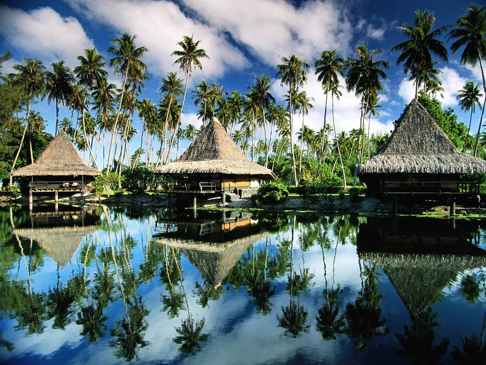 Three thatched-roof bungalows stand on stilts over calm water, surrounded by palm trees, with reflections in the water.
