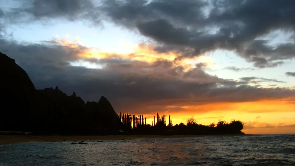 A dramatic sunset over a tropical coastline with silhouetted mountains and dark clouds.