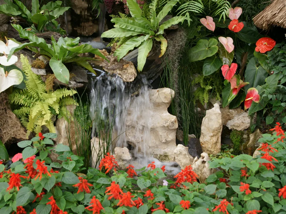 A small waterfall cascades over rocks in a lush garden filled with tropical plants and red flowers.