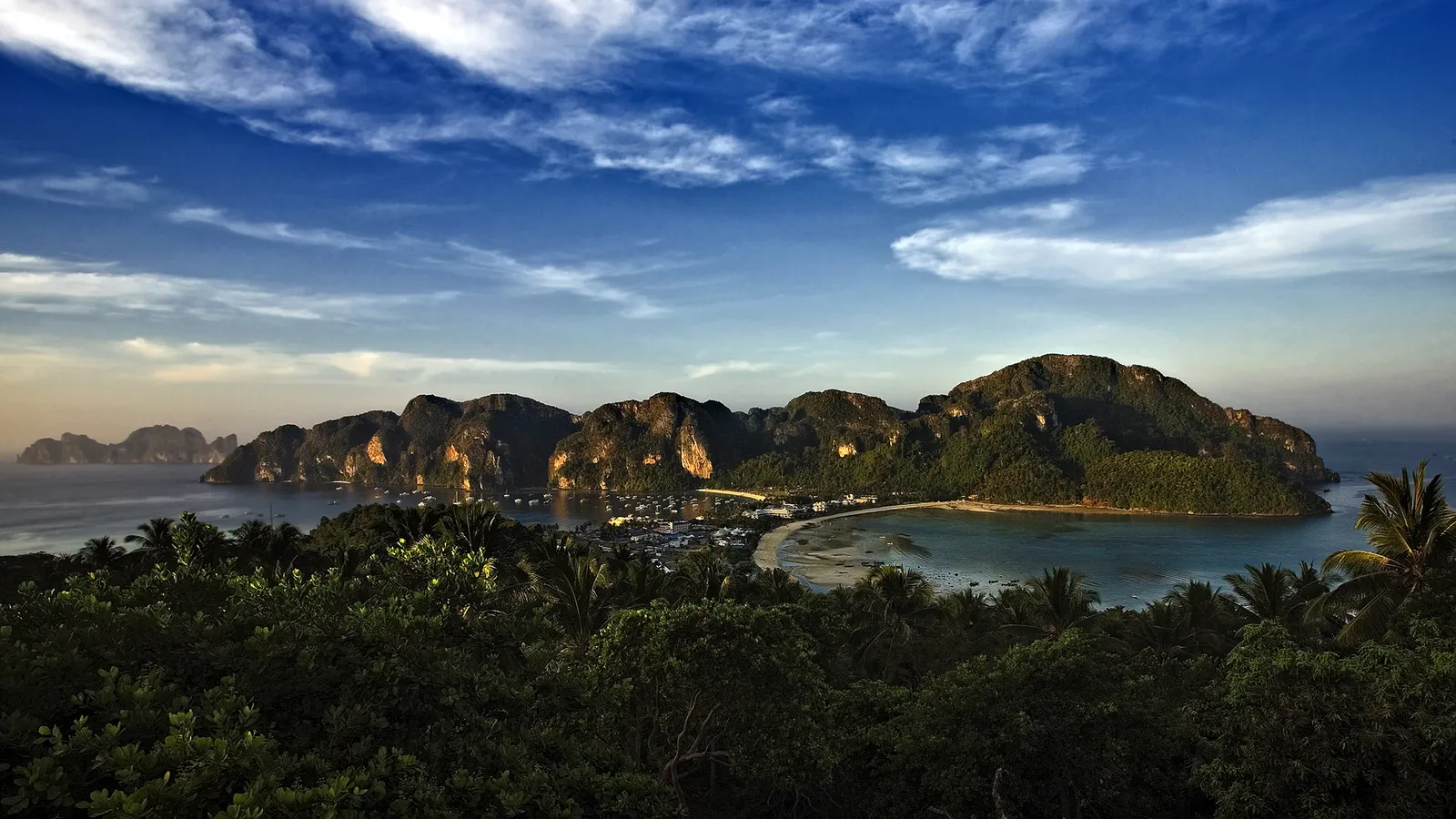 A scenic view of a tropical island bay with lush green cliffs and a sandy beach, under a blue sky with clouds.