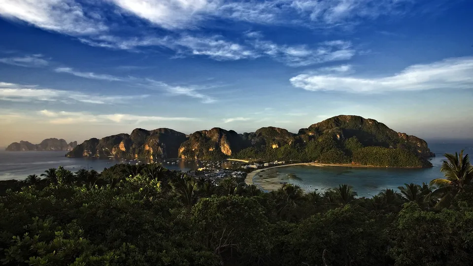 A scenic view of a tropical island bay with lush green cliffs and a sandy beach, under a blue sky with clouds.