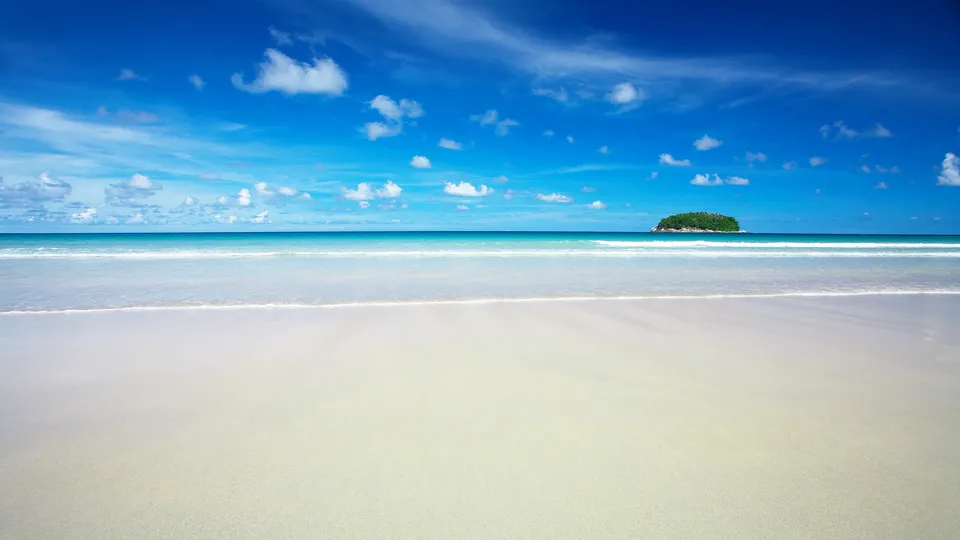 A wide, sandy beach meets the turquoise ocean under a bright blue sky with scattered white clouds. A small, green island is visible on the horizon.