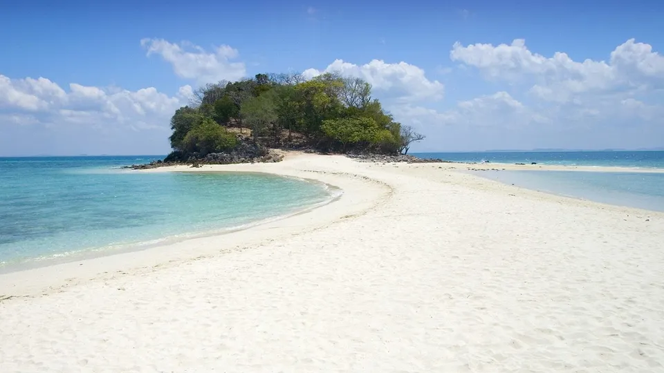 A curved white sand beach leads to a small, tree-covered island in clear turquoise ocean water under a blue sky with white clouds.