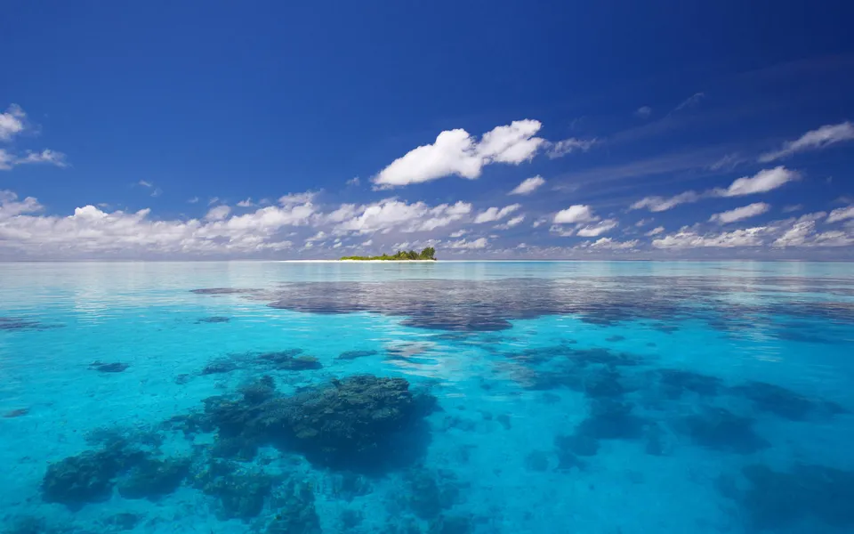 A small, lush green island sits on the horizon in a vast expanse of clear turquoise ocean under a bright blue sky with scattered white clouds.