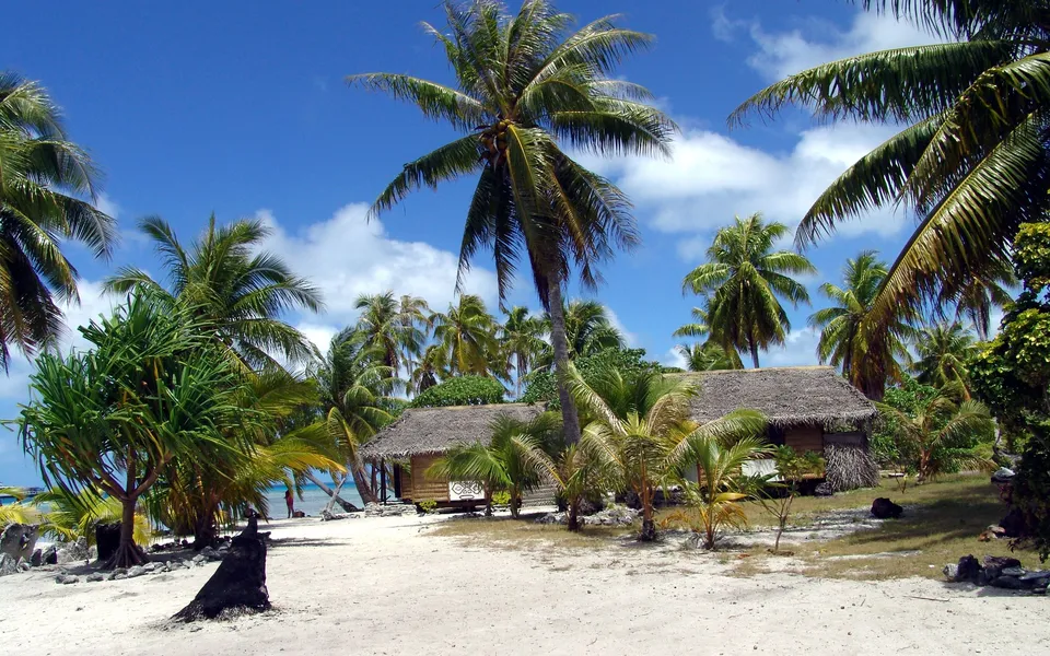 Thatched roof bungalows nestled among palm trees on a sandy beach with a bright blue sky.
