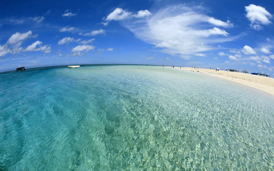A wide shot of a tropical island beach with crystal clear turquoise water and white sand under a blue sky with clouds.