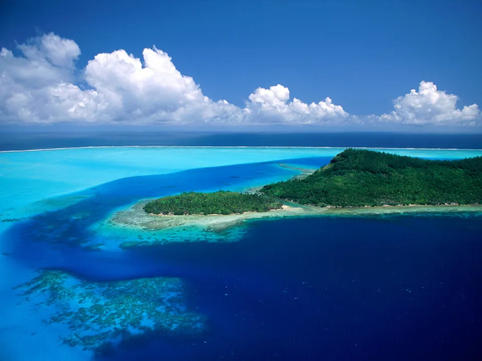 Aerial view of a lush green island surrounded by vibrant turquoise and deep blue ocean waters under a bright blue sky with white clouds.
