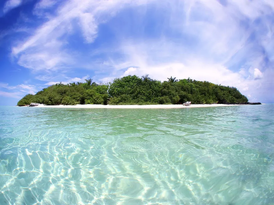 A lush green tropical island with a white sand beach is surrounded by crystal clear turquoise water under a bright blue sky with wispy clouds.