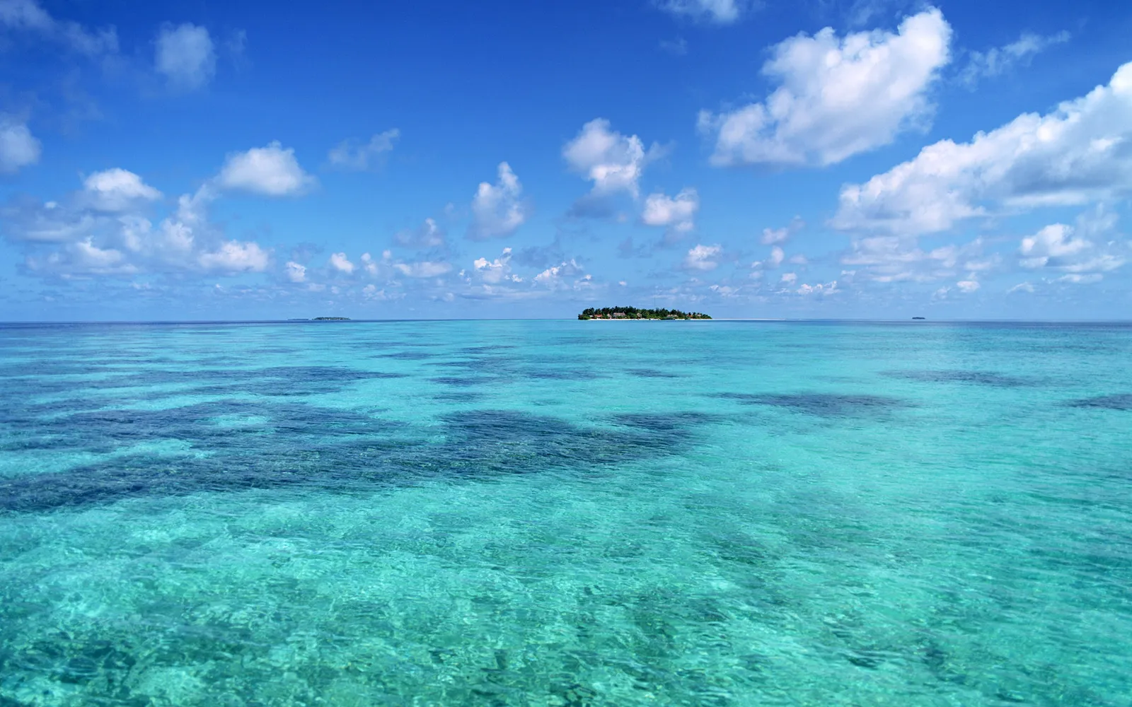 A small tropical island with buildings is visible in the distance across clear turquoise ocean water under a blue sky with white clouds.