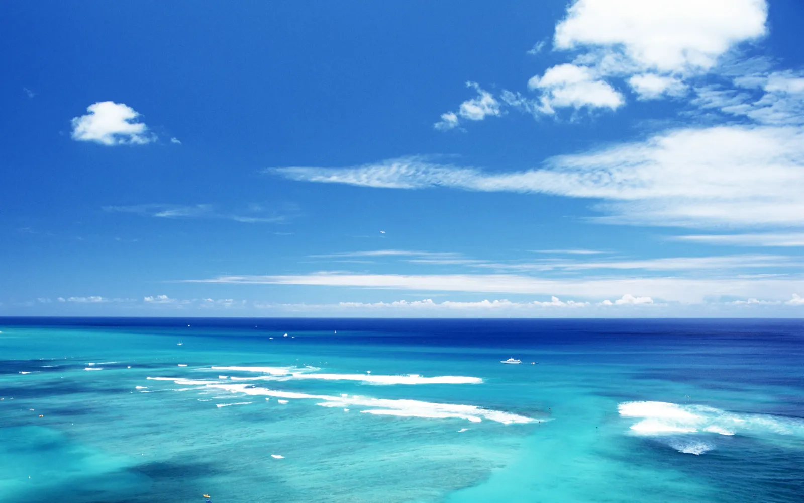 An aerial view of a tropical ocean with vibrant turquoise waters and white waves under a clear blue sky with scattered clouds.