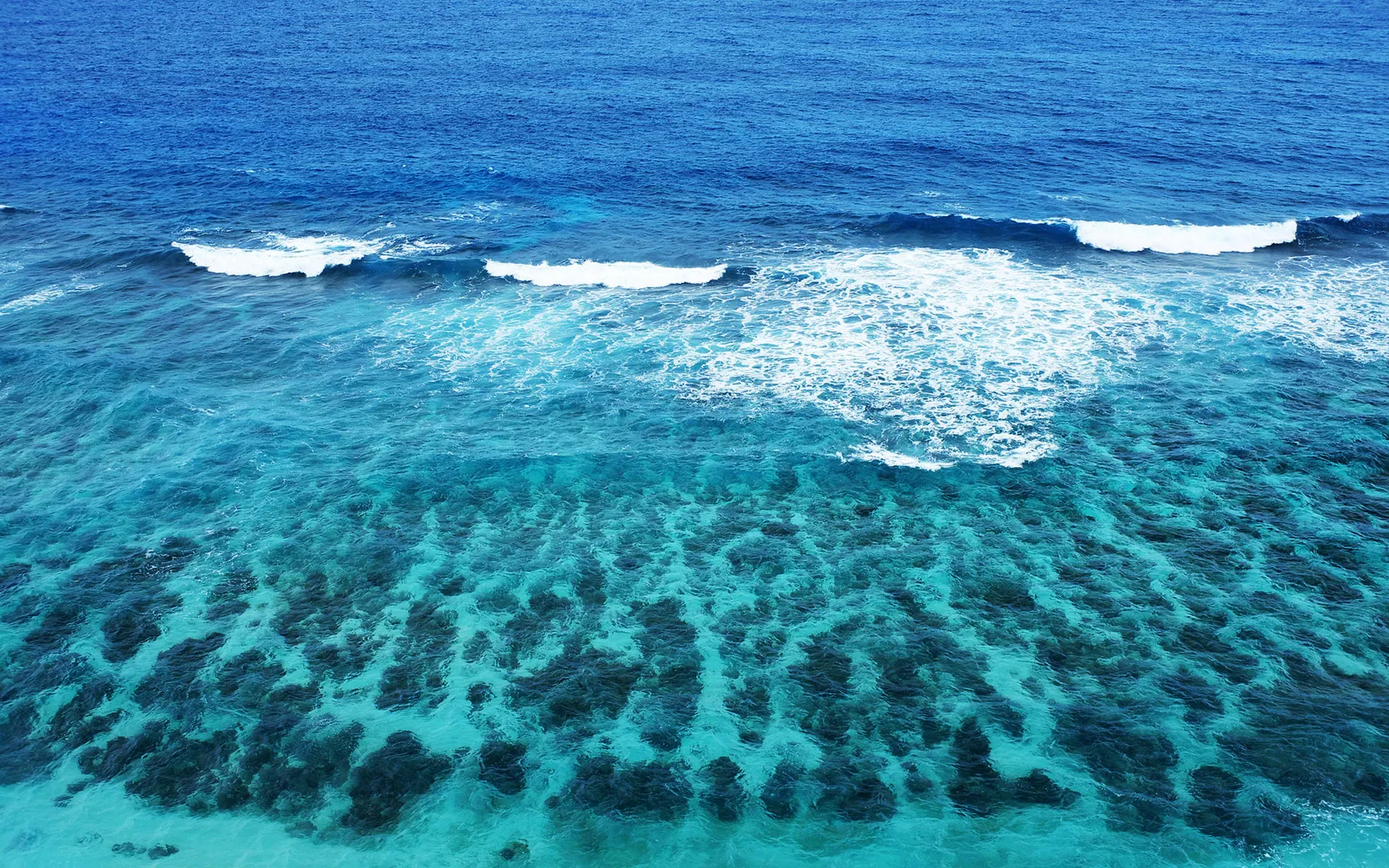 Aerial view of clear turquoise ocean water revealing a vibrant coral reef below with gentle waves breaking.