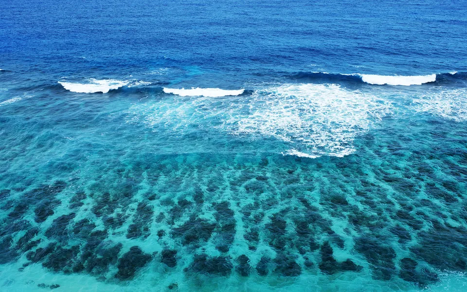 Aerial view of clear turquoise ocean water revealing a vibrant coral reef below with gentle waves breaking.