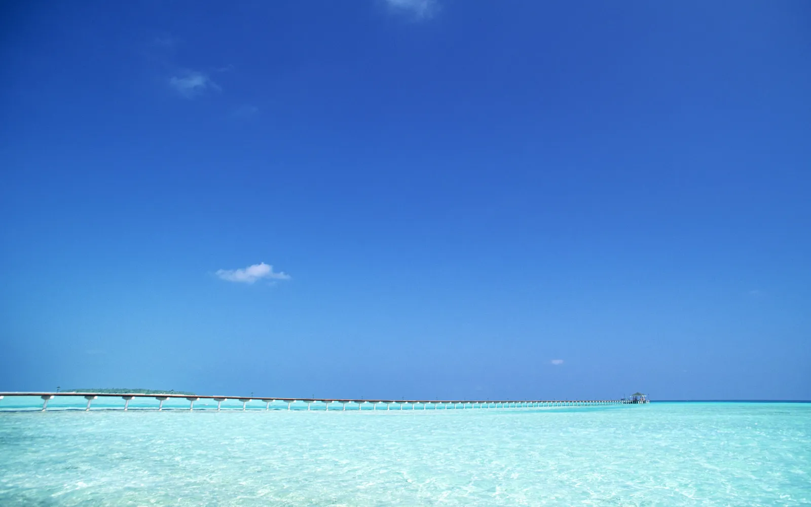A long pier stretches across clear turquoise water under a bright blue sky with a few small clouds.