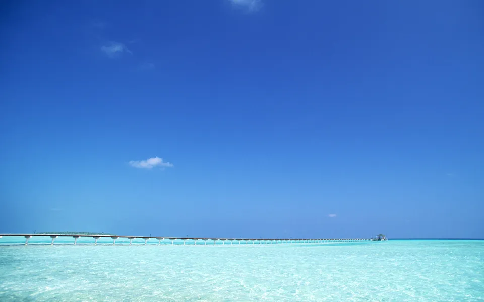 A long pier stretches across clear turquoise water under a bright blue sky with a few small clouds.