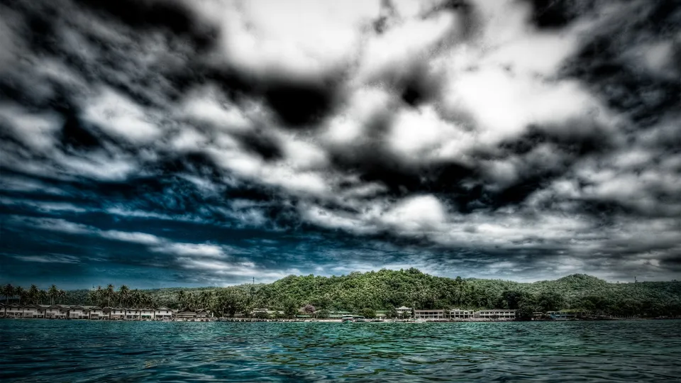 A tropical resort with white buildings and palm trees sits on a lush green island under a dramatic, dark, cloudy sky.