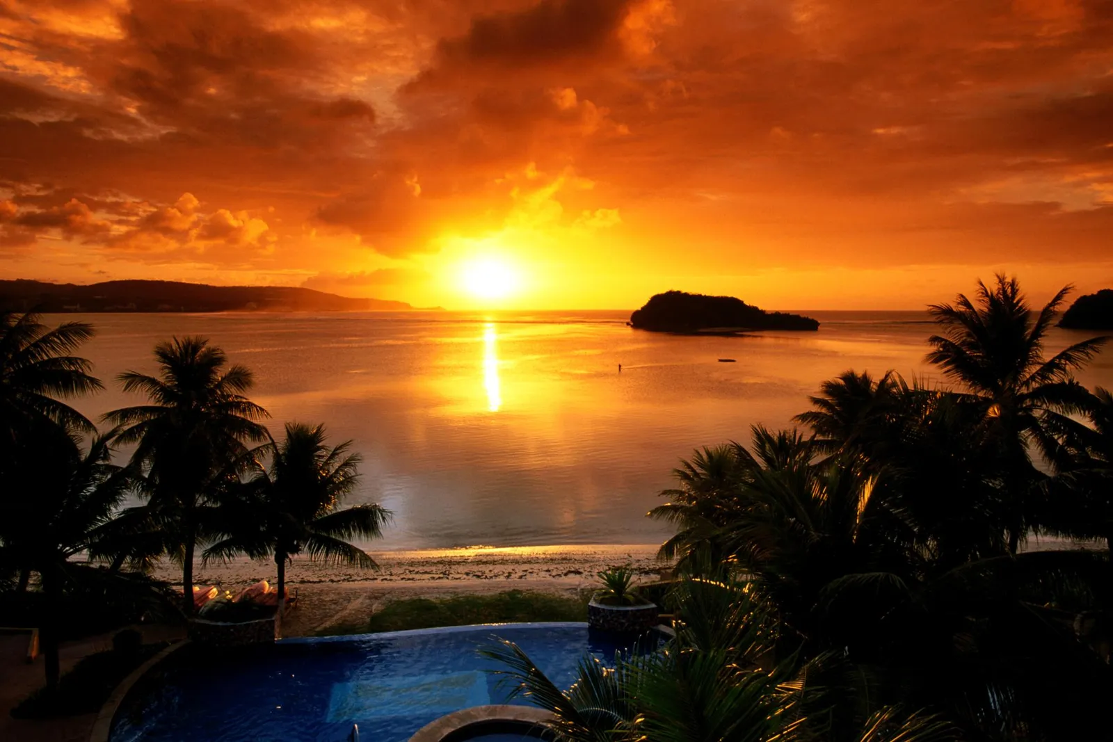 A vibrant orange and yellow sunset over a calm bay, with silhouetted palm trees in the foreground and a small island in the distance.