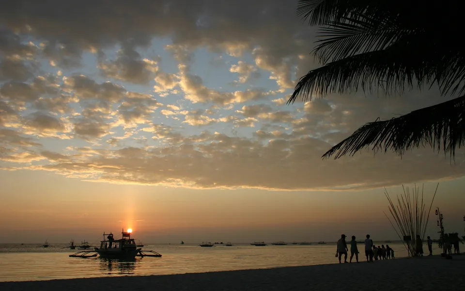 A vibrant sunset over the ocean with silhouetted boats and people on the beach, framed by palm fronds.