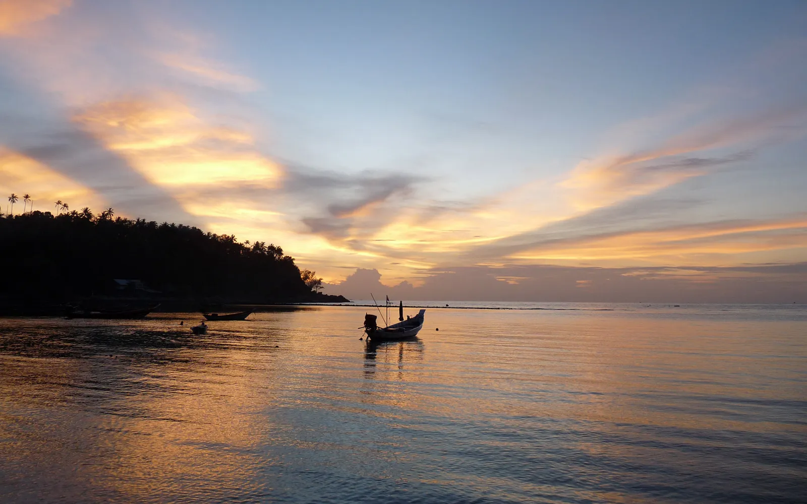 A lone fishing boat floats on calm water during a colorful sunset with silhouetted palm trees on the shore.