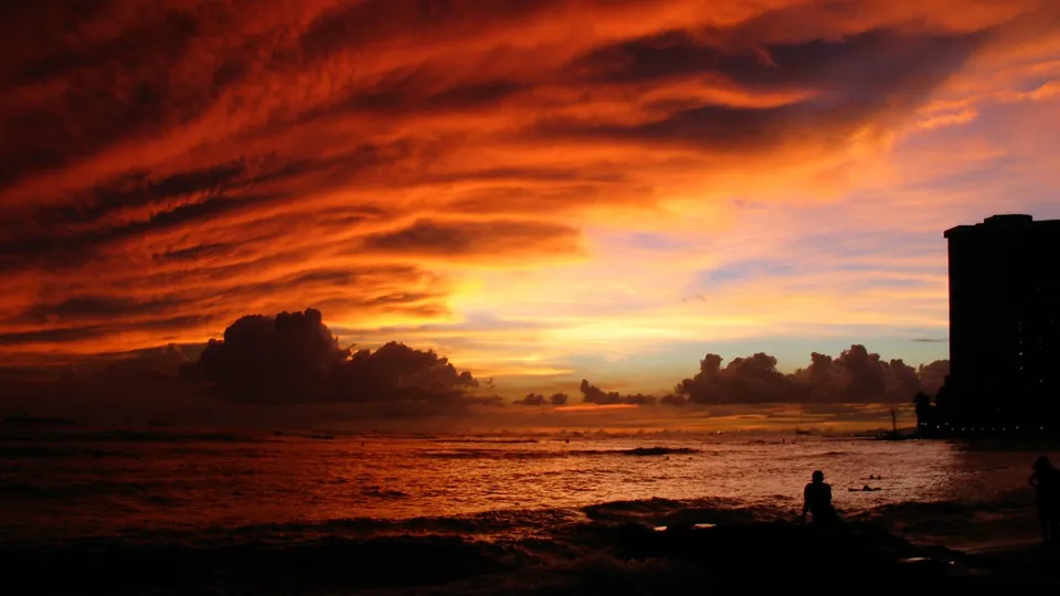 A vibrant sunset with fiery orange and red clouds over a dark ocean with silhouetted figures on the shore.