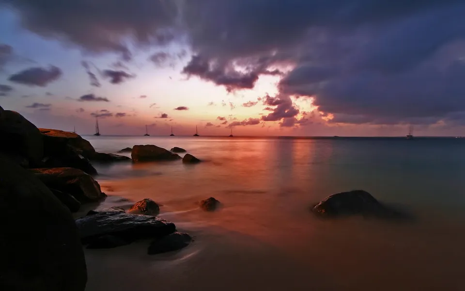 A dramatic sunset over the ocean with sailboats silhouetted on the horizon and rocks in the foreground.