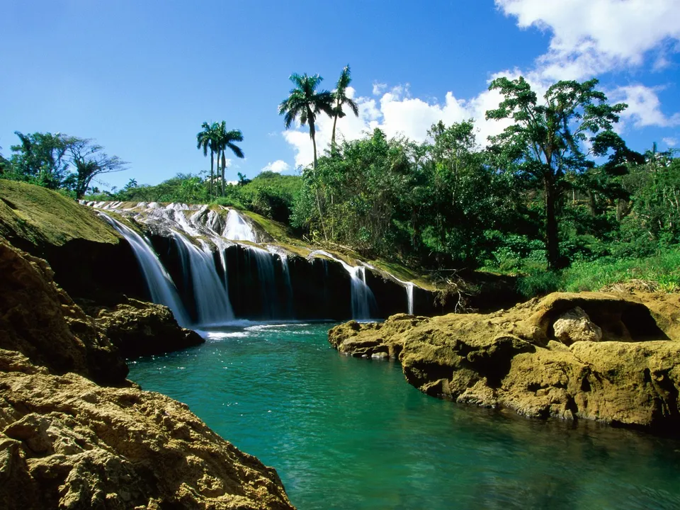 A wide, multi-tiered waterfall flows over mossy rocks into a turquoise pool, surrounded by dense green trees and palm trees under a blue sky.