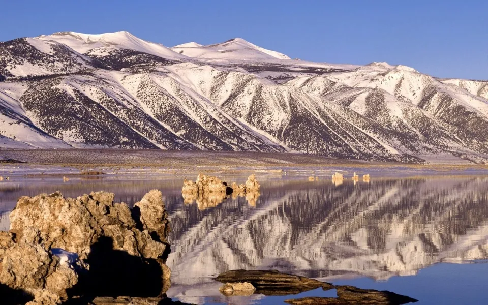 Tufa rocks in a lake reflecting snow-dusted mountains under a blue sky.