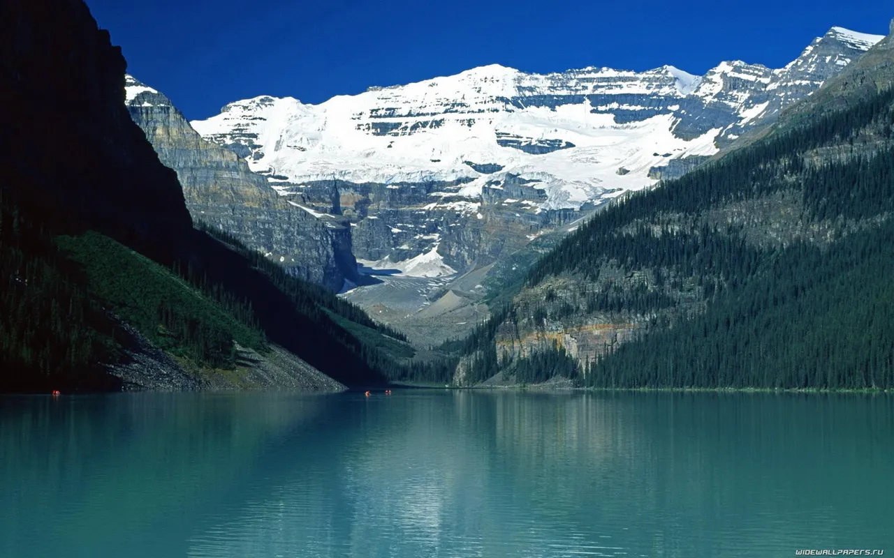 A wide, calm turquoise lake reflects snow-capped mountains and green forested slopes under a clear blue sky.