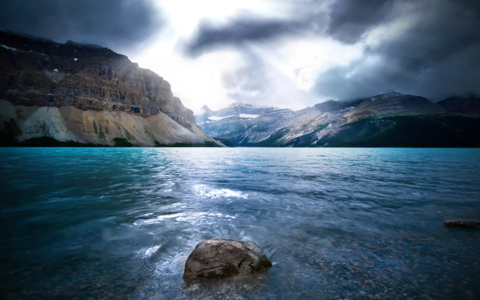 A wide, turquoise lake with mountains in the background under a dramatic, cloudy sky with sunbeams breaking through.