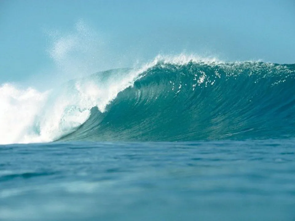 A powerful turquoise ocean wave breaks with white foam under a clear, pale blue sky.