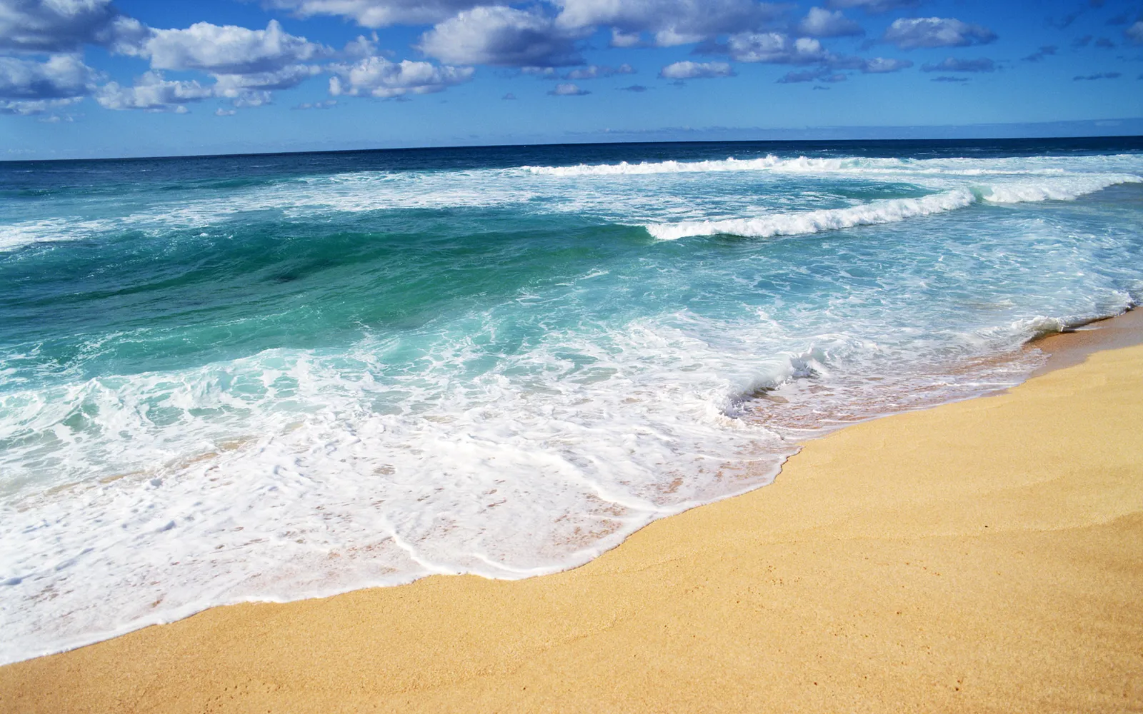 Turquoise ocean waves roll onto a golden sand beach under a blue sky with scattered clouds.