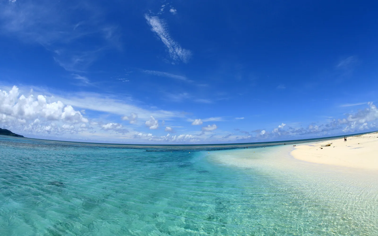 A panoramic view of a tropical beach with crystal clear turquoise water and white sand under a bright blue sky with scattered clouds.